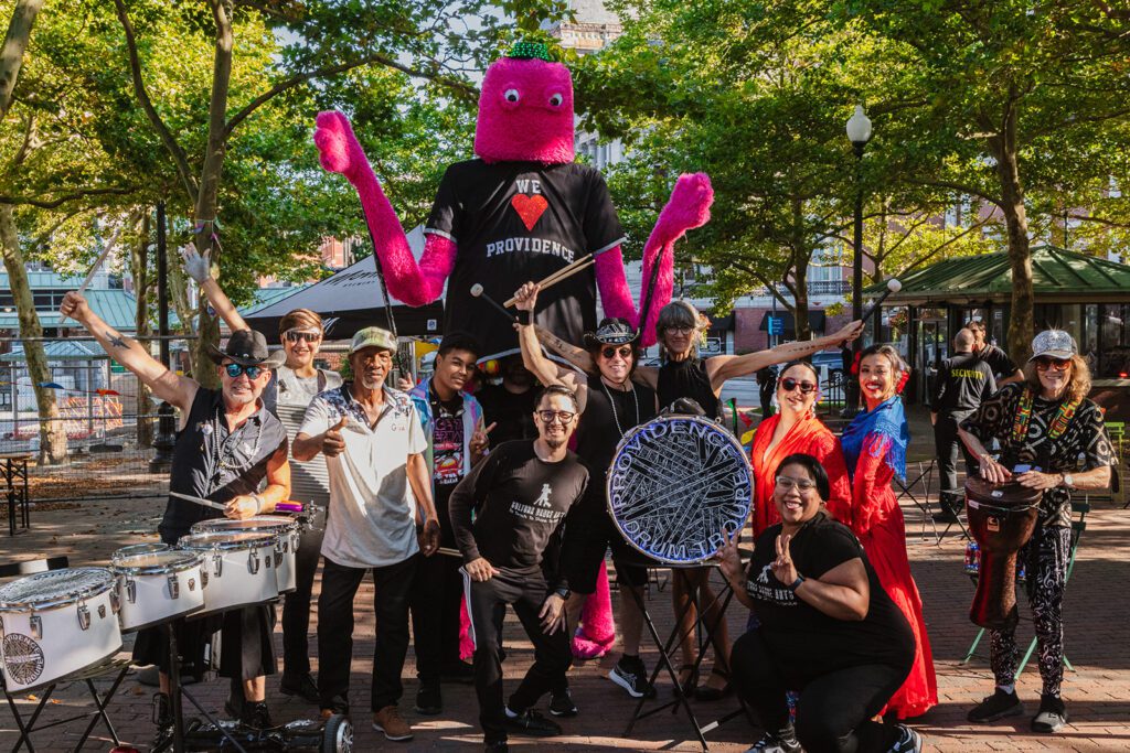 Group photo of the Providence Drum Troupe with colorful performers, musicians, and a large pink octopus mascot wearing a ‘We Love Providence’ shirt, celebrating community art and culture in a vibrant outdoor park setting.