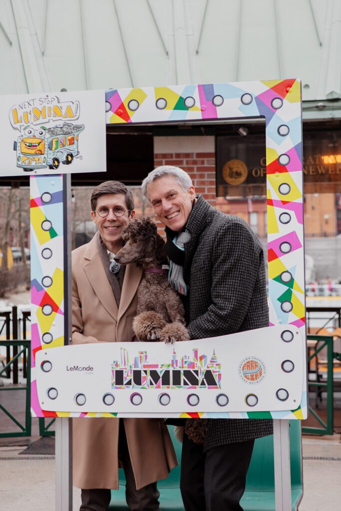 Providence Mayor Brett Smiley and his husband Jim DeRentis with their dog at the LUMINA festival, framed by a colorful sign.