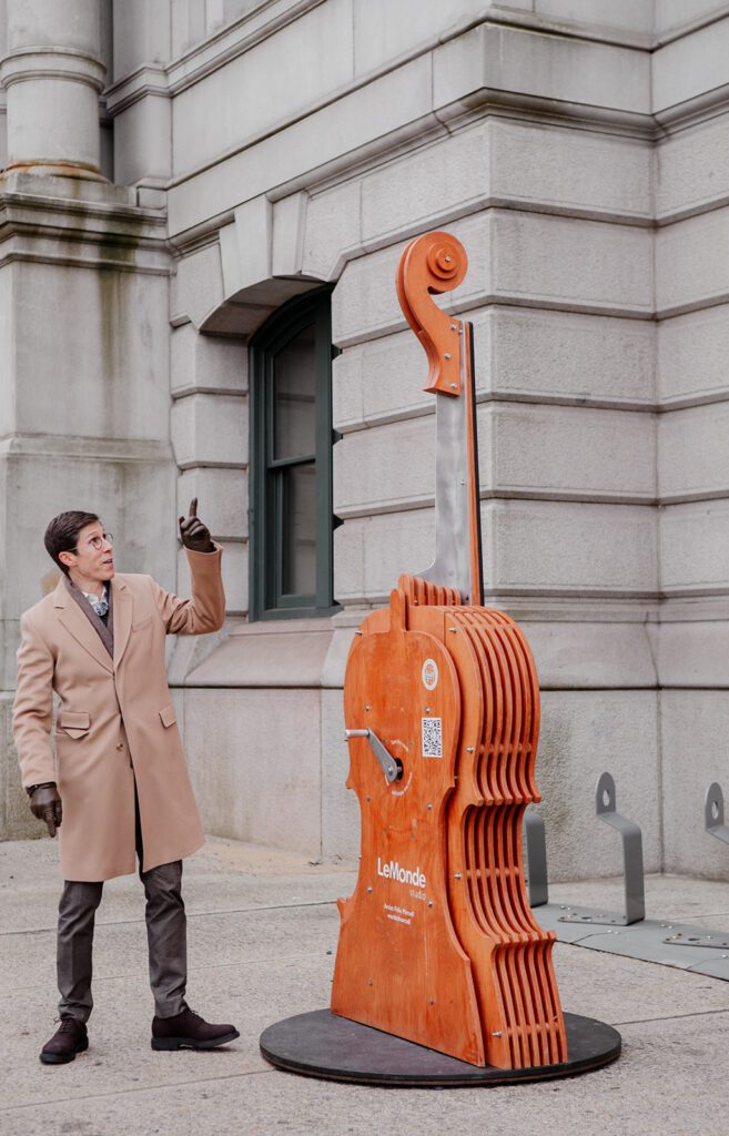 Providence Mayor Brett Smiley gestures toward the Harmonies Violin sculpture by Le Monde Studio, outside a stone building.