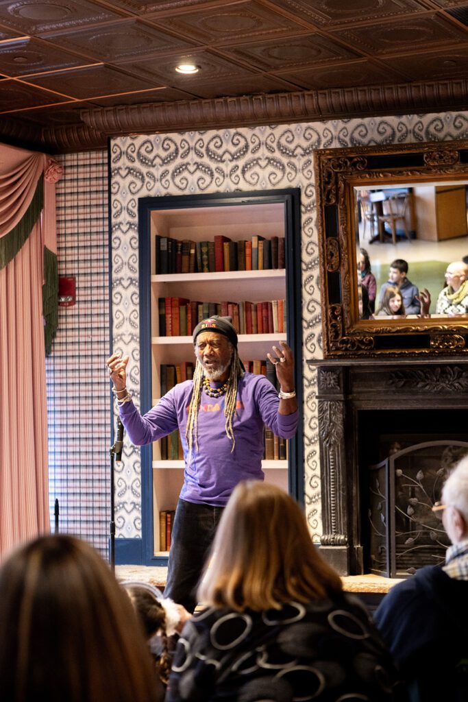 Len Cabral storytelling in a cozy room with bookshelves and a decorative fireplace, engaging an audience.