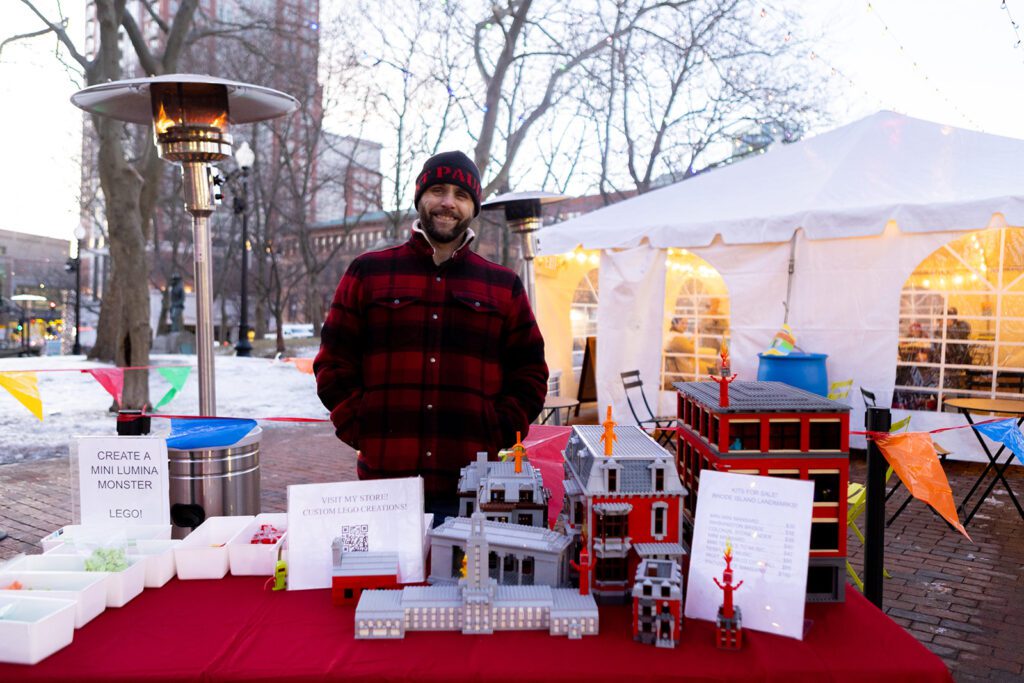 Rhode Island LEGO Artist Andrew Grover stands at an outdoor booth displaying custom LEGO creations, with a festive tent in the background.