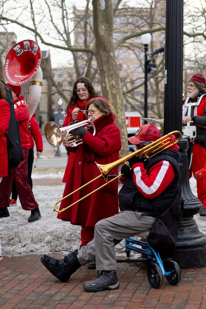 Members of the Extraordinary Rendition Band perform outdoors, wearing red outfits and playing brass instruments.