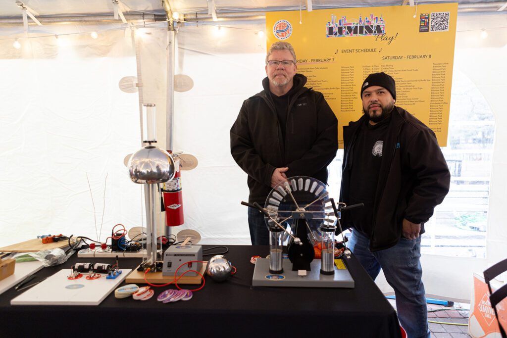 Two men stand by a table with electrical science equipment under a tent, with an event schedule in the background.