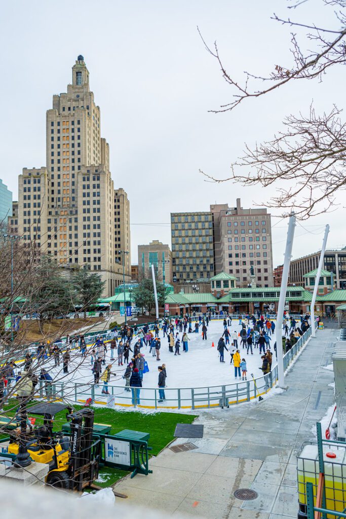 Providence Rink with skaters, surrounded by tall buildings, and a clear sky.