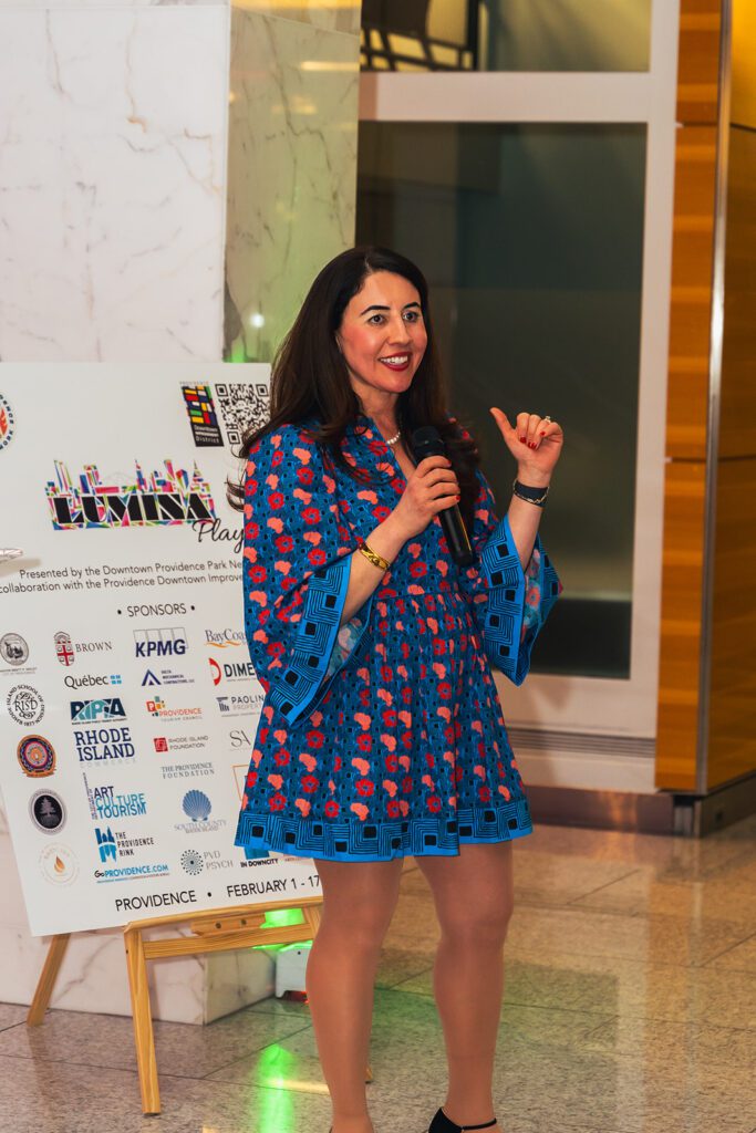 Downtown Providence Parks Network Executive Director Nora Barré speaking at the LUMINA VIP event, holding a microphone, with a promotional poster in the background.