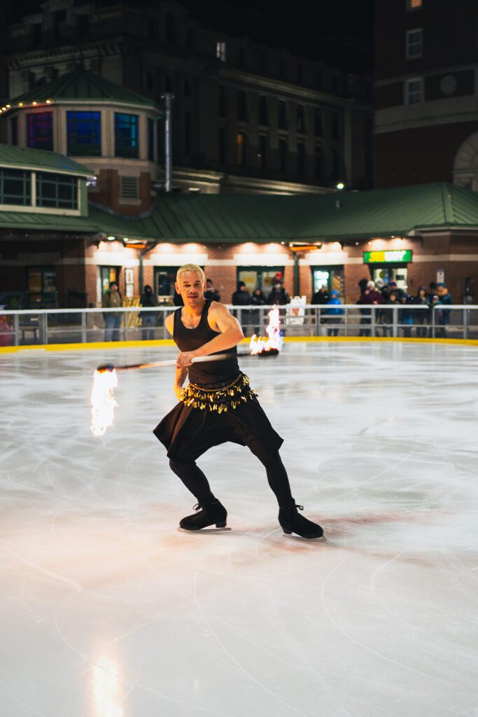 Performer with flaming batons on ice at night, part of Ice Dance International, with a backdrop of a lit building and spectators.