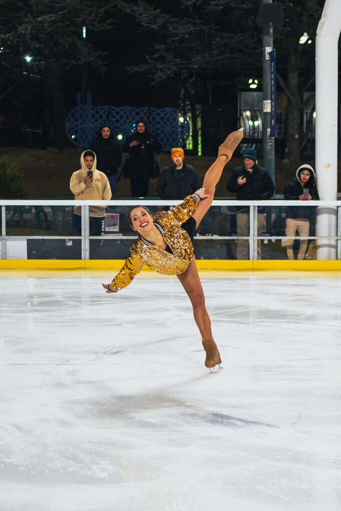 A figure skater from "Ice Dance International" performs a graceful spiral on the rink, wearing a sparkling gold outfit.