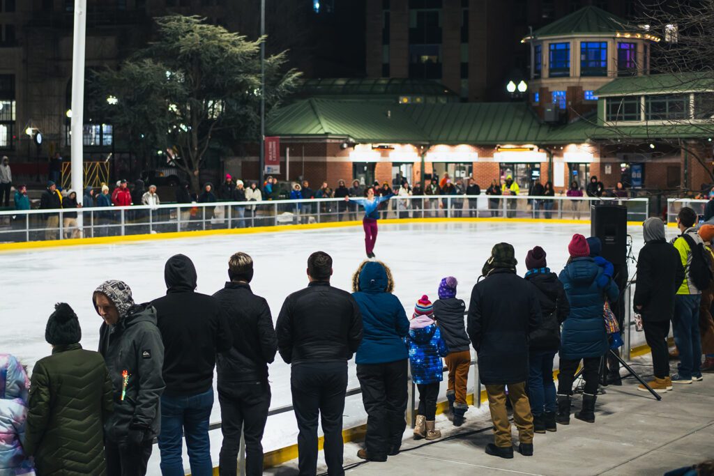 Audience watching an "Ice Dance International" performance at an outdoor rink, surrounded by lights and buildings at night.