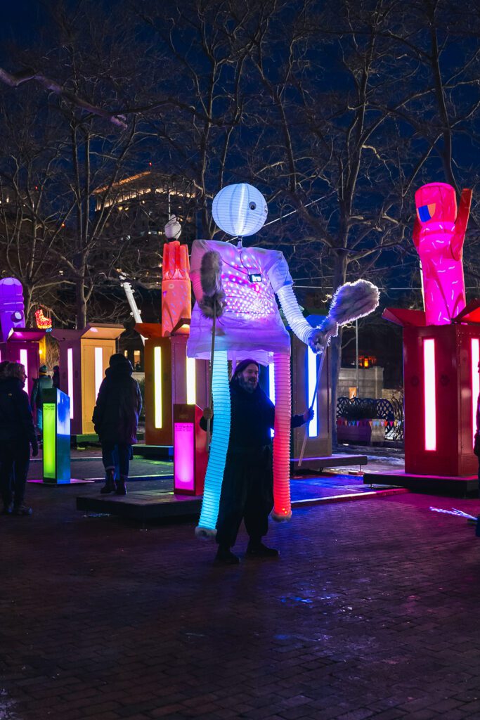 Colorful illuminated puppet and people at a nighttime outdoor festival with vibrant lights and artistic installations.