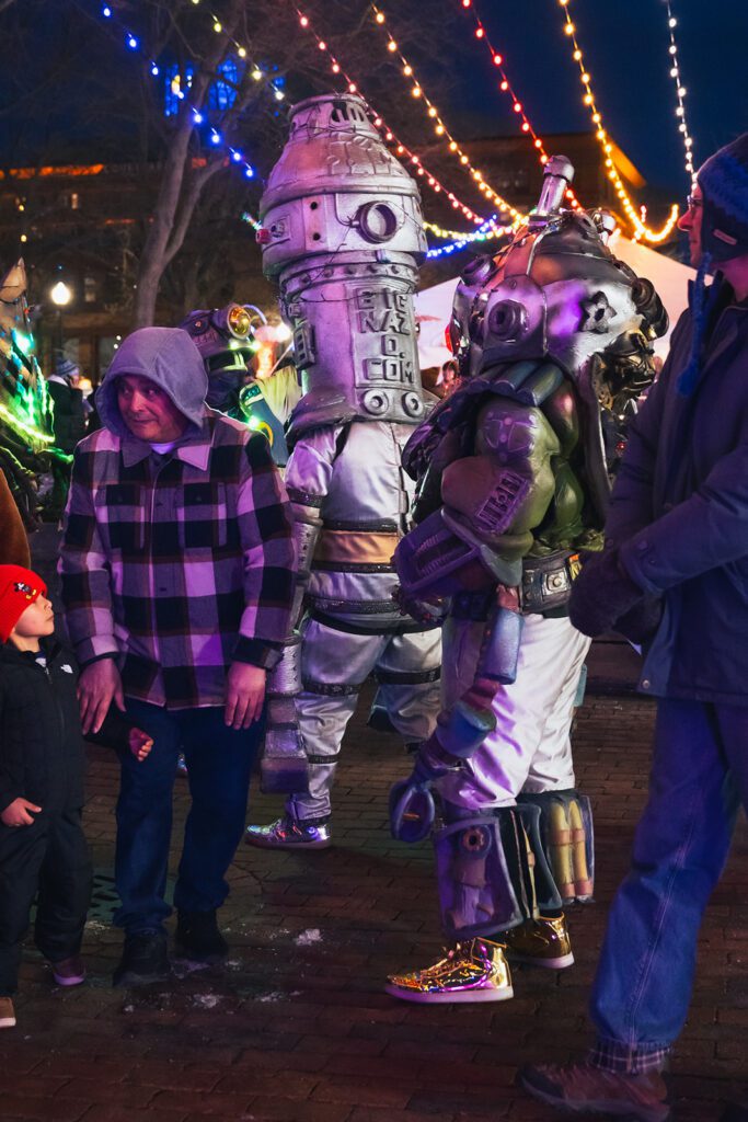 Big NAZO performers in futuristic costumes interact with festival-goers under colorful string lights at an outdoor event.