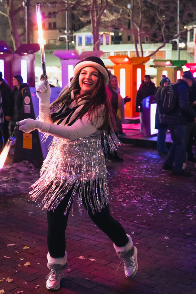 Woman in a sparkly outfit joyfully twirling a light stick at a vibrant nighttime festival.