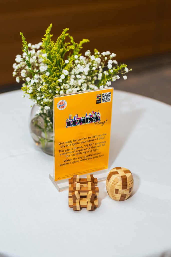 Event flyer on table with wooden puzzles and flowers, promoting Lumina's "PLAY" theme and city light-up experience.