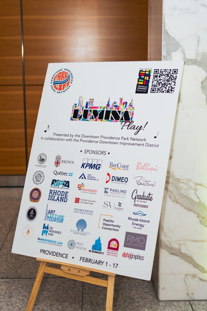 Event poster with colorful "LUMINA Play!" logo, sponsors, and QR code, displayed on an easel in Providence, February 1-17.