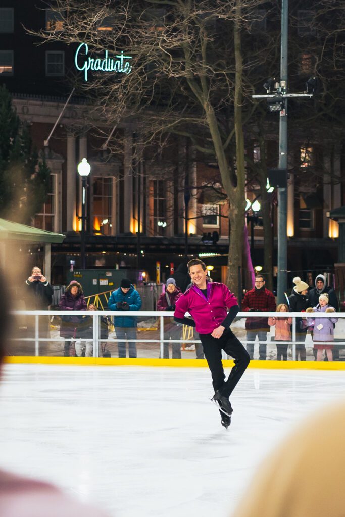 Skater performing at Ice Dance International event, wearing a magenta shirt. Spectators watch near the "Graduate" hotel sign.