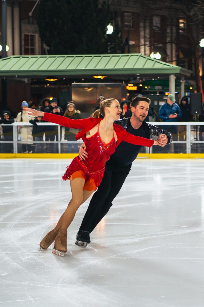 Figure skaters perform an elegant routine on an outdoor rink during an "Ice Dance International" event, with onlookers in the background.