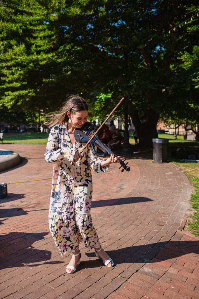 Andrea's Bowstring performing a lively violin piece outdoors, wearing a flowing patterned dress, with Biltmore Park as the backdrop.