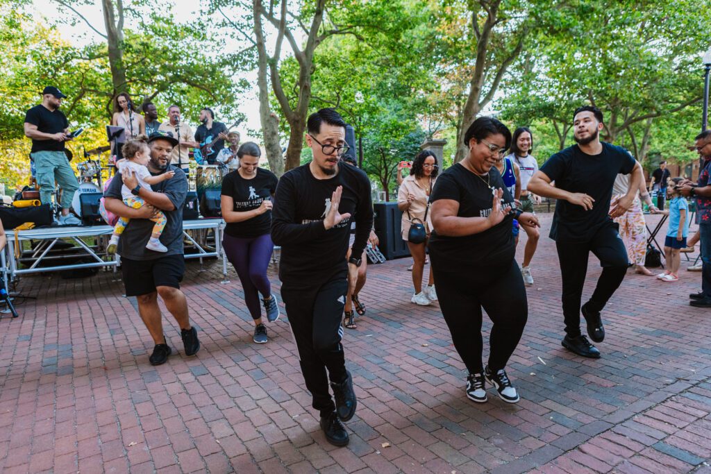 Cultura Dance Arts group performing an energetic dance routine in Biltmore Park, accompanied by a live band on stage, surrounded by a lively crowd under a canopy of trees.