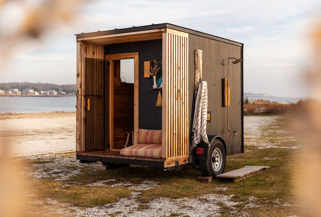 Altaer Space Mobile Sauna featuring a sleek black exterior with wood accents, open to reveal a cozy interior, set on a scenic lakeside location with serene views in the background.