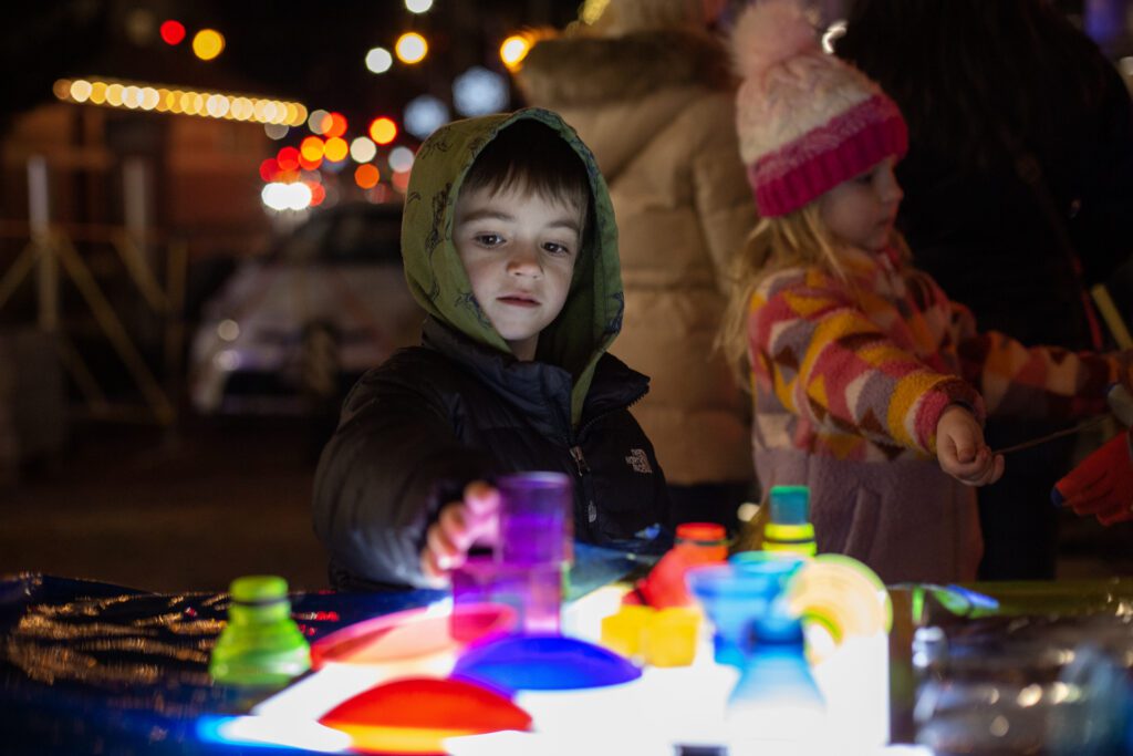 Children engaging in the ‘It’s Lit!’ light projection activity by P3, featuring vibrant illuminated shapes on a table, with a cozy nighttime setting and festive background lighting.