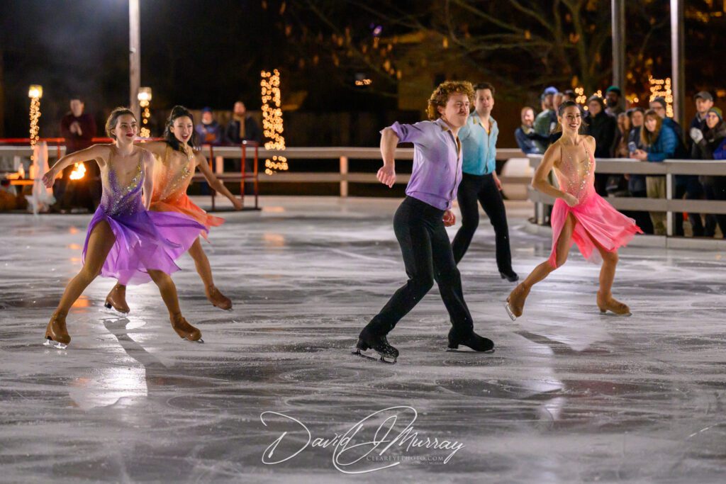 Ice Dance International ensemble performing a synchronized routine on an outdoor ice rink, featuring skaters in vibrant costumes with a festive, illuminated nighttime setting in the background.