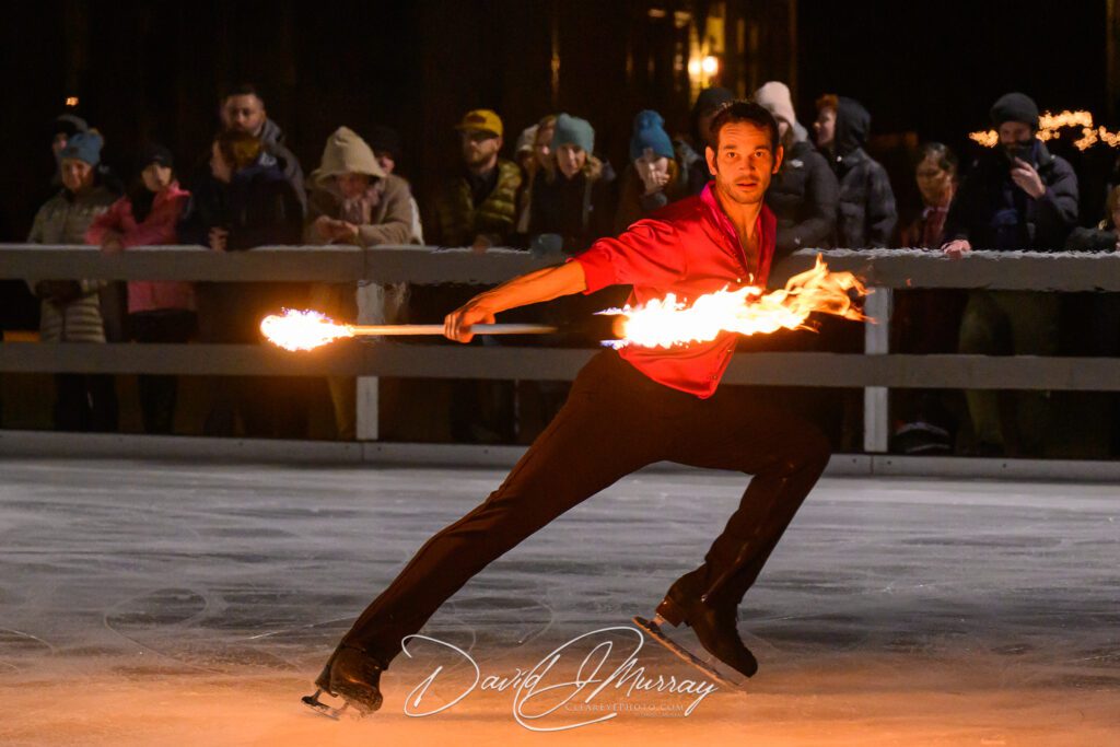 Ice Dance International performance featuring a skater in a dynamic pose on ice, wielding a flaming baton, with a captivated audience in the background during a nighttime show.