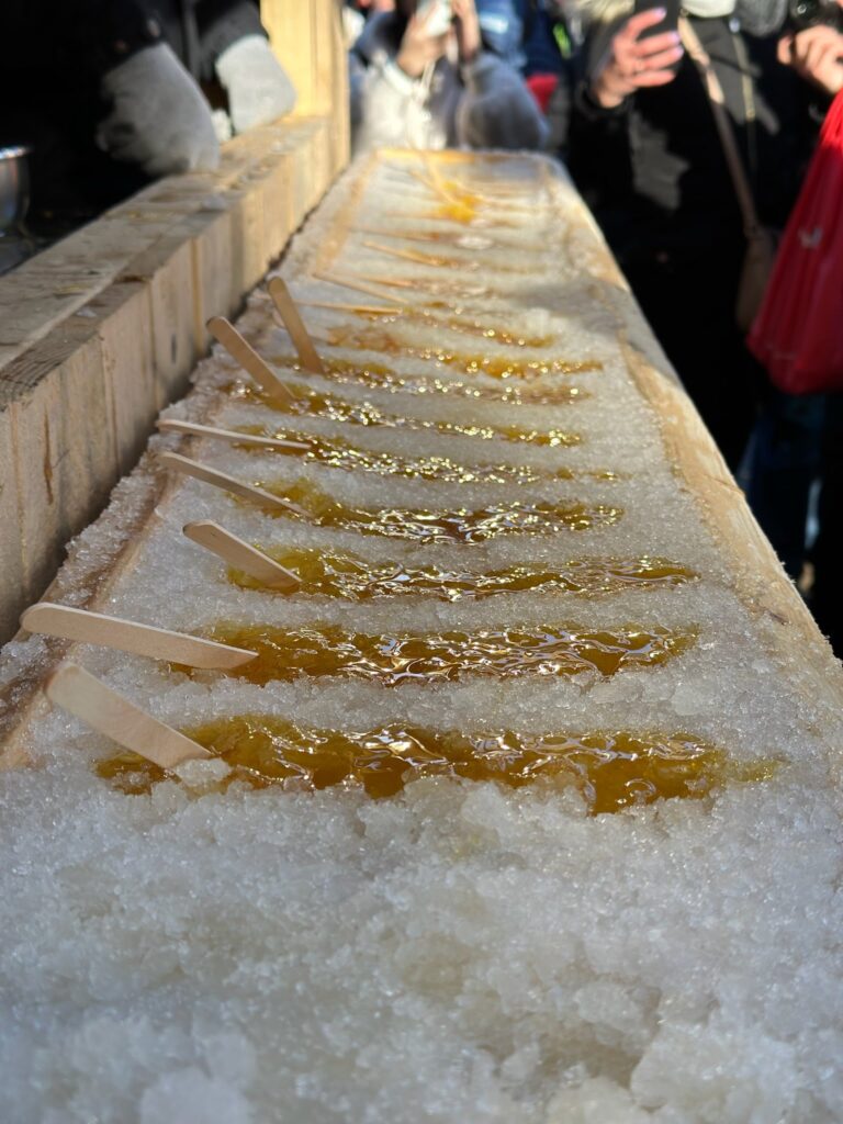 Maple taffy on snow setup featuring wooden sticks laid on fresh snow with golden maple syrup hardening into candy, surrounded by onlookers enjoying the traditional treat.