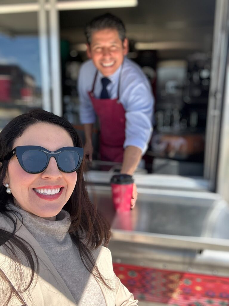 Nora Barré enjoying a visit to Café Modesto, smiling in the foreground with a barista in a red apron handing out a coffee cup from the café counter in the background.”