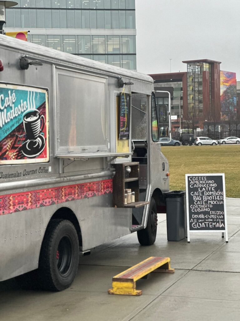 Café Modesto’s gourmet coffee truck parked at the Providence Pedestrian Bridge, featuring vibrant artwork on its side and a chalkboard menu listing specialty coffee options.