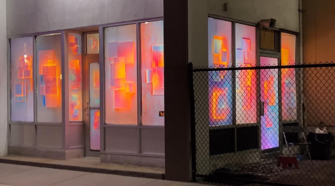 A storefront window installation illuminated at night with vibrant pink, orange, and blue geometric light projections, creating a dynamic and colorful display.