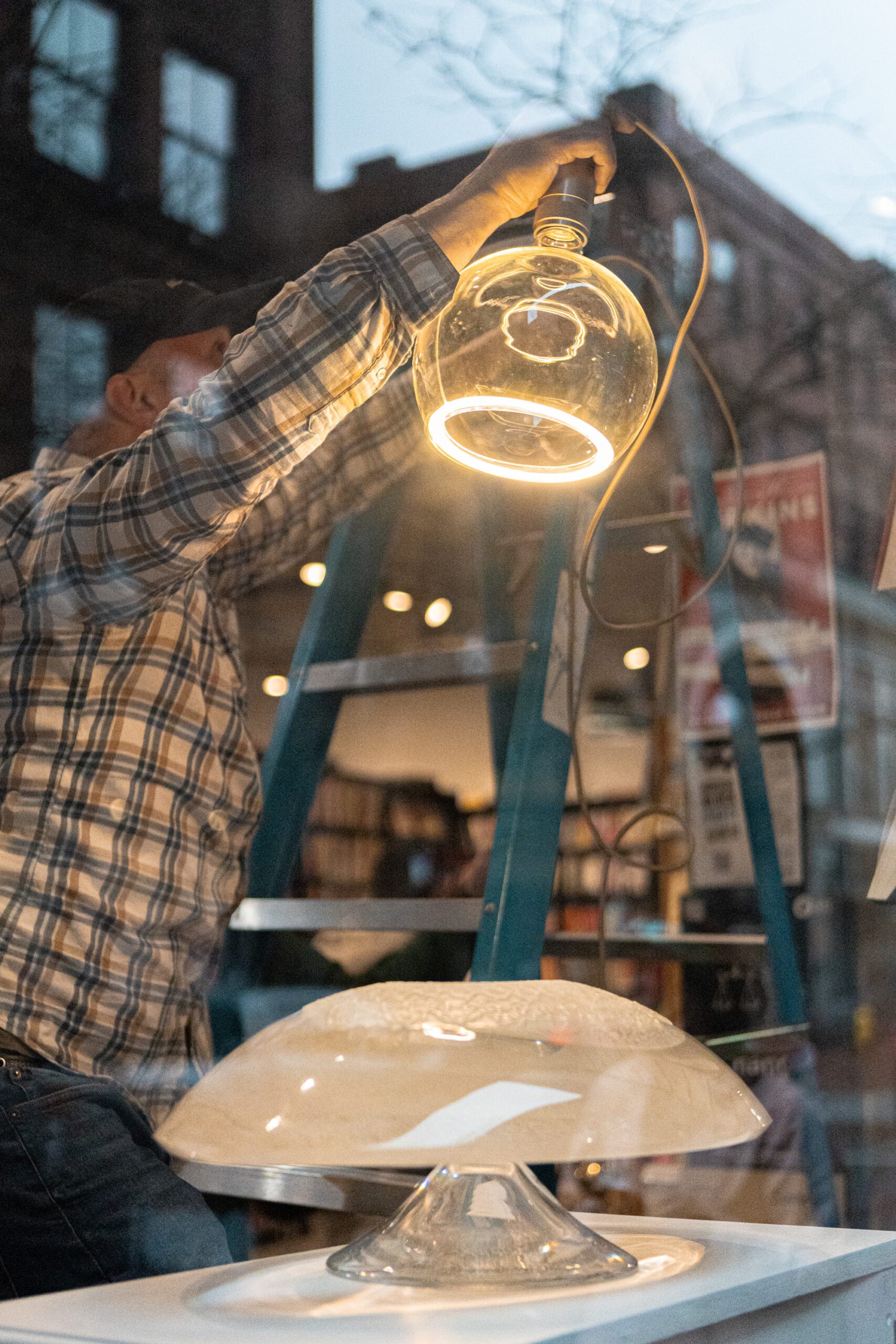 A man in a plaid shirt installs a glowing glass pendant light in a storefront window, with a blue ladder and a blurred background of bookshelves and city reflections. A sculptural glass piece sits on a white display table below.