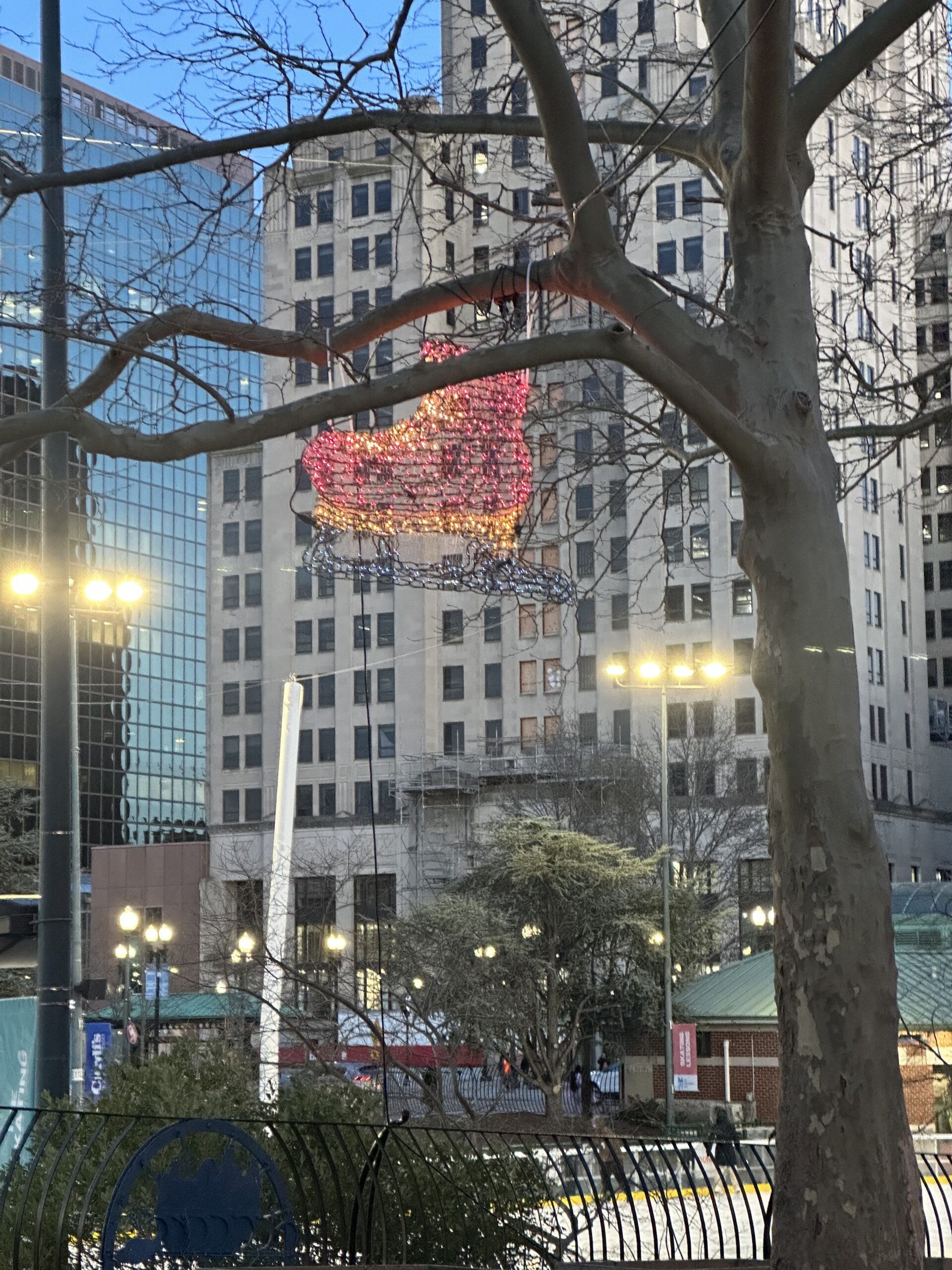 A festive illuminated decoration of a red and gold ice skate hangs from tree branches in an urban park during early evening. Tall buildings with a mix of glass and stone facades form the background, while streetlights cast a warm glow over the scene. A skating rink and a green-roofed pavilion are visible in the lower portion of the image.