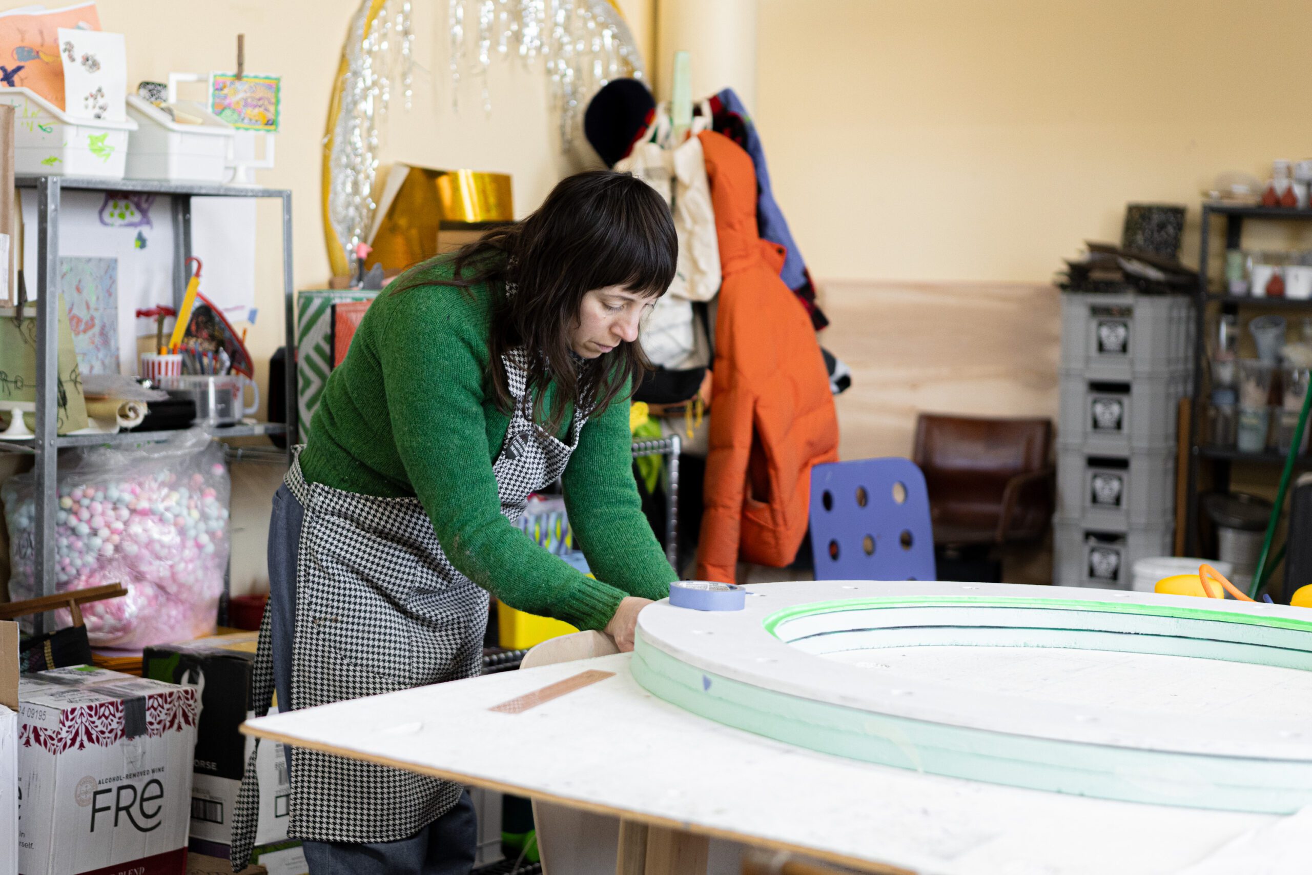 An artist wearing a green sweater and checkered apron works on a circular foam structure in a creative studio filled with art supplies, tools, and colorful decorations.