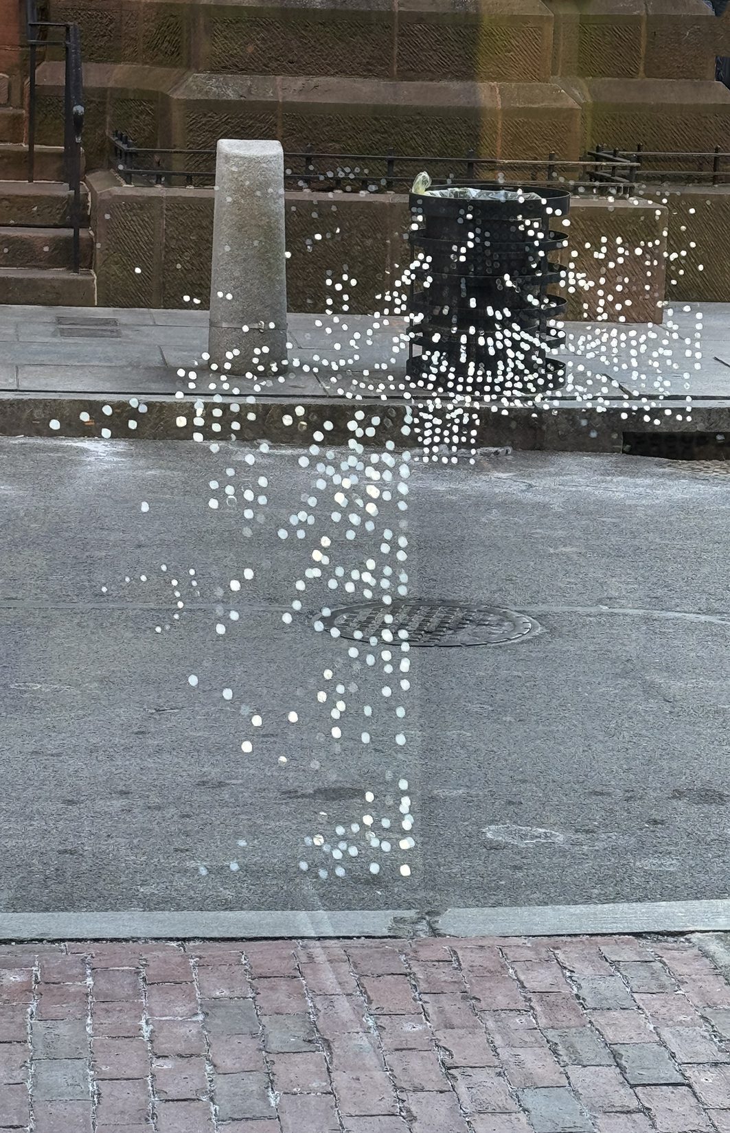 A glass window with a pattern of white dots reflects the urban street outside, blending the dots into the scene of a sidewalk, a bollard, a trash can, and a historic stone building in the background.